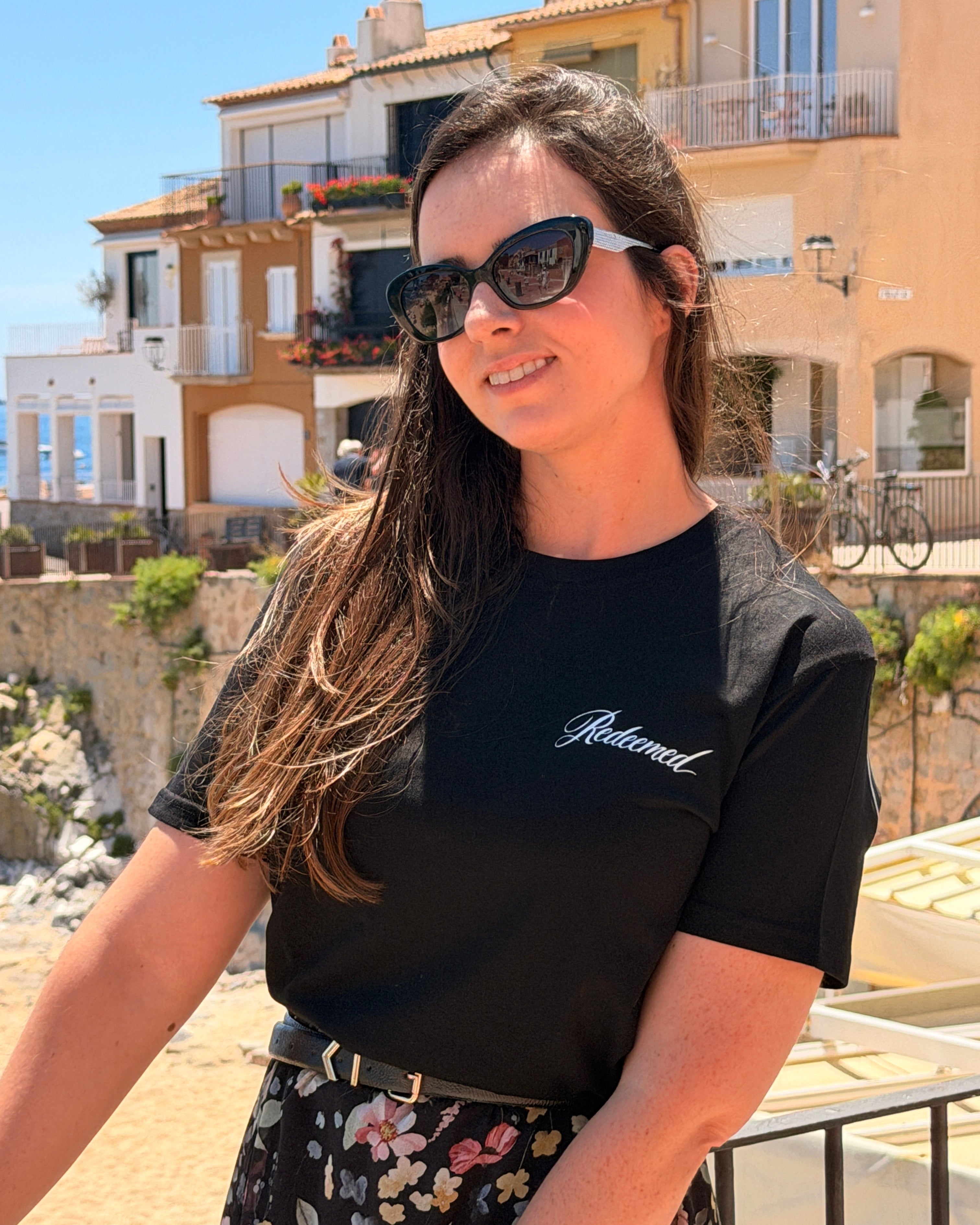 A woman wearing black cat-eye sunglasses and a black t-shirt with 'Relational' written in white script on the chest, posing outdoors in front of coastal buildings.