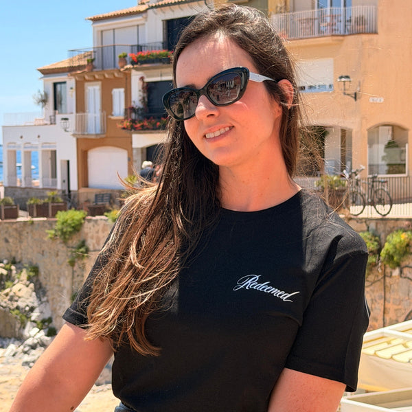 A woman wearing black cat-eye sunglasses and a black t-shirt with 'Relational' written in white script on the chest, posing outdoors in front of coastal buildings.