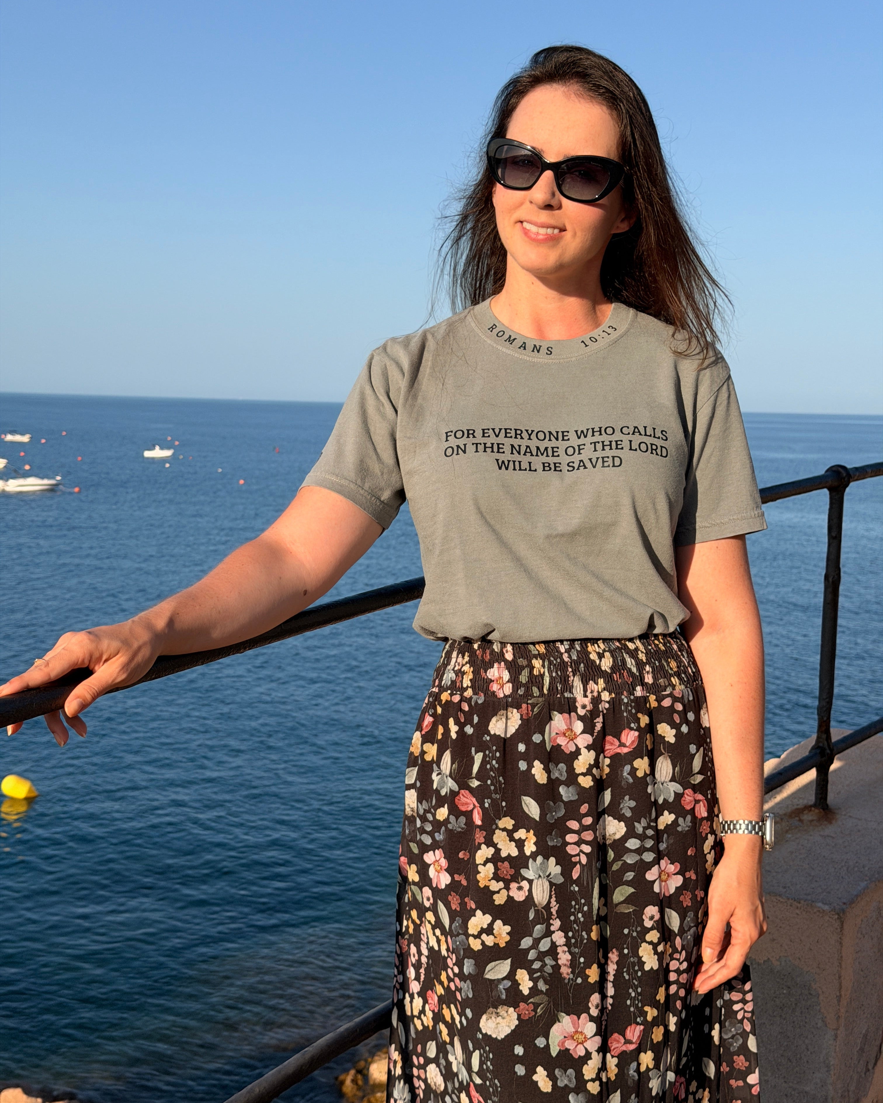 A woman wearing a gray graphic t-shirt and a black floral maxi skirt stands on a balcony overlooking the blue sea and sky. The t-shirt features the text 'FOR EVERYONE WHO CALLS ON THE NAME OF THE LORD WILL BE SAVIED'.