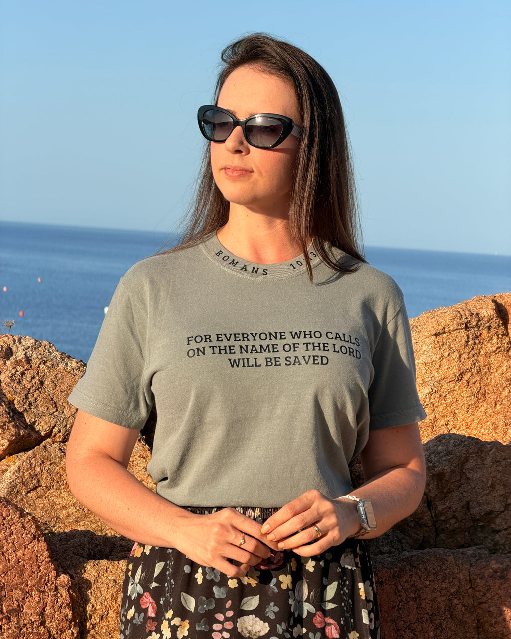 A woman wearing a sage green graphic t-shirt with black text reading 'FOR EVERYONE WHO CALLS ON THE NAME OF THE LORD WILL BE SAVED' and a floral skirt, standing outdoors against a rocky shoreline and blue sky.