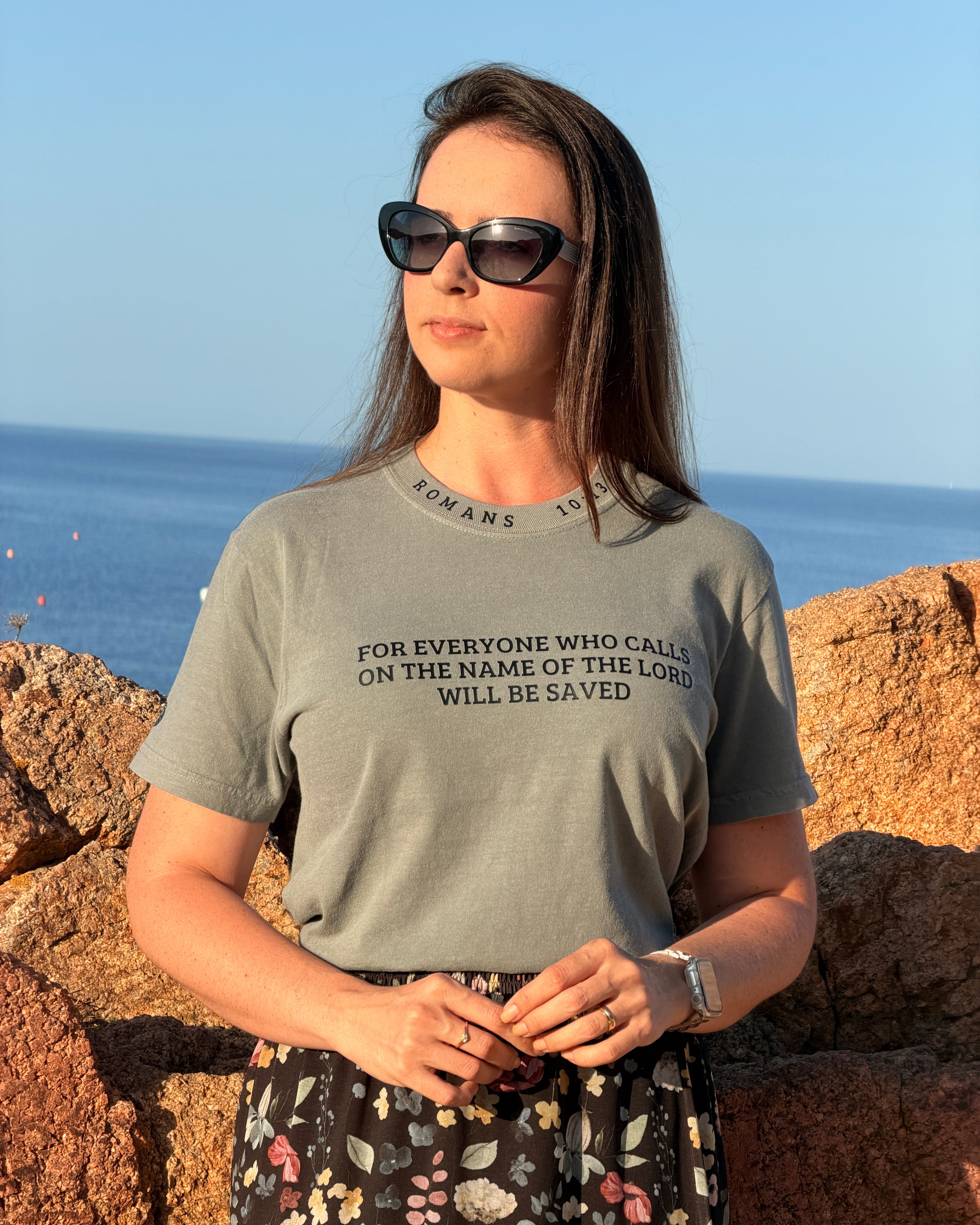 A woman wearing a sage green graphic t-shirt with black text reading 'FOR EVERYONE WHO CALLS ON THE NAME OF THE LORD WILL BE SAVED' and a floral skirt, standing outdoors against a rocky shoreline and blue sky.