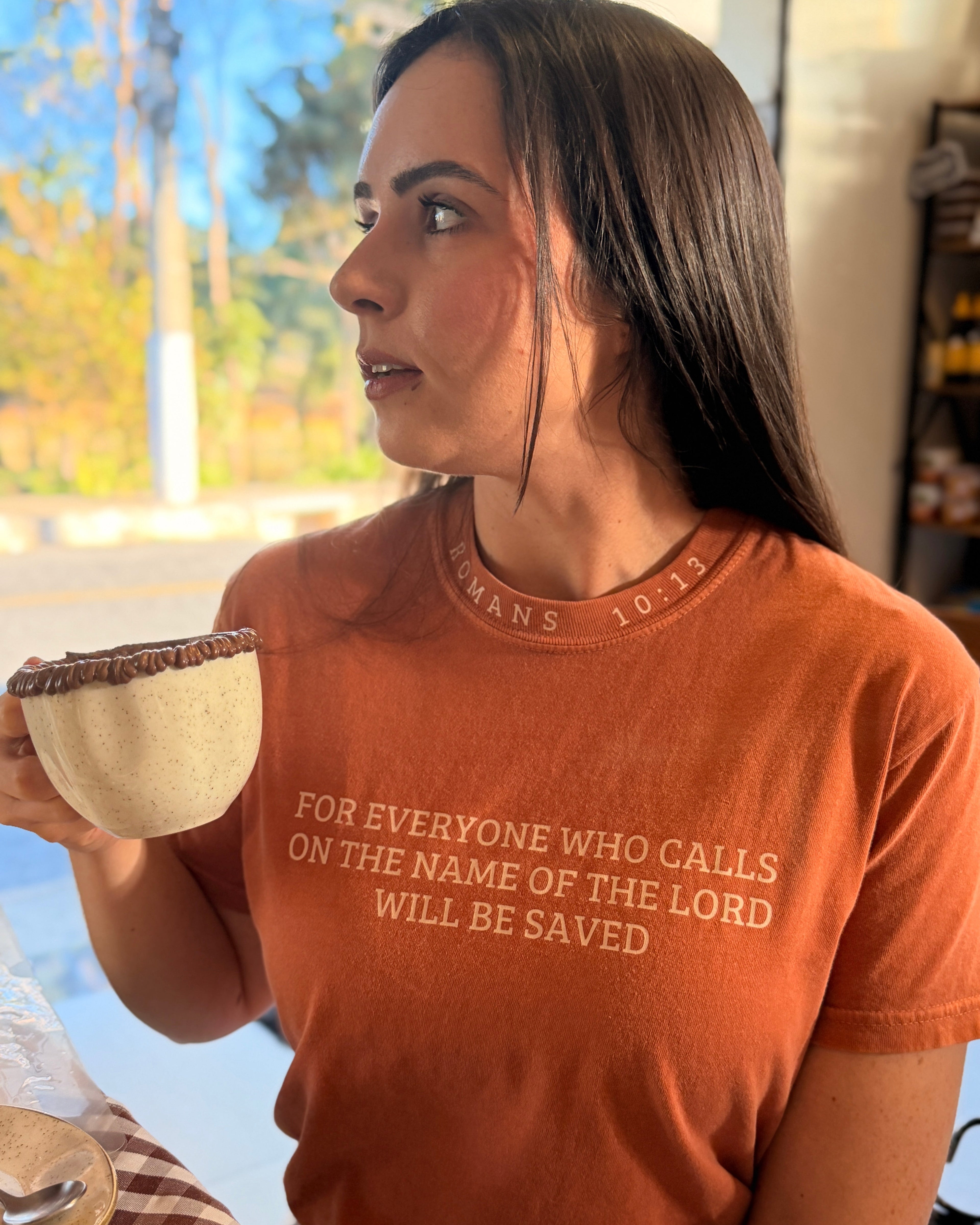 A woman wearing an orange crewneck t-shirt with a white printed Bible verse, holding a small ceramic bowl in a brightly lit indoor setting.