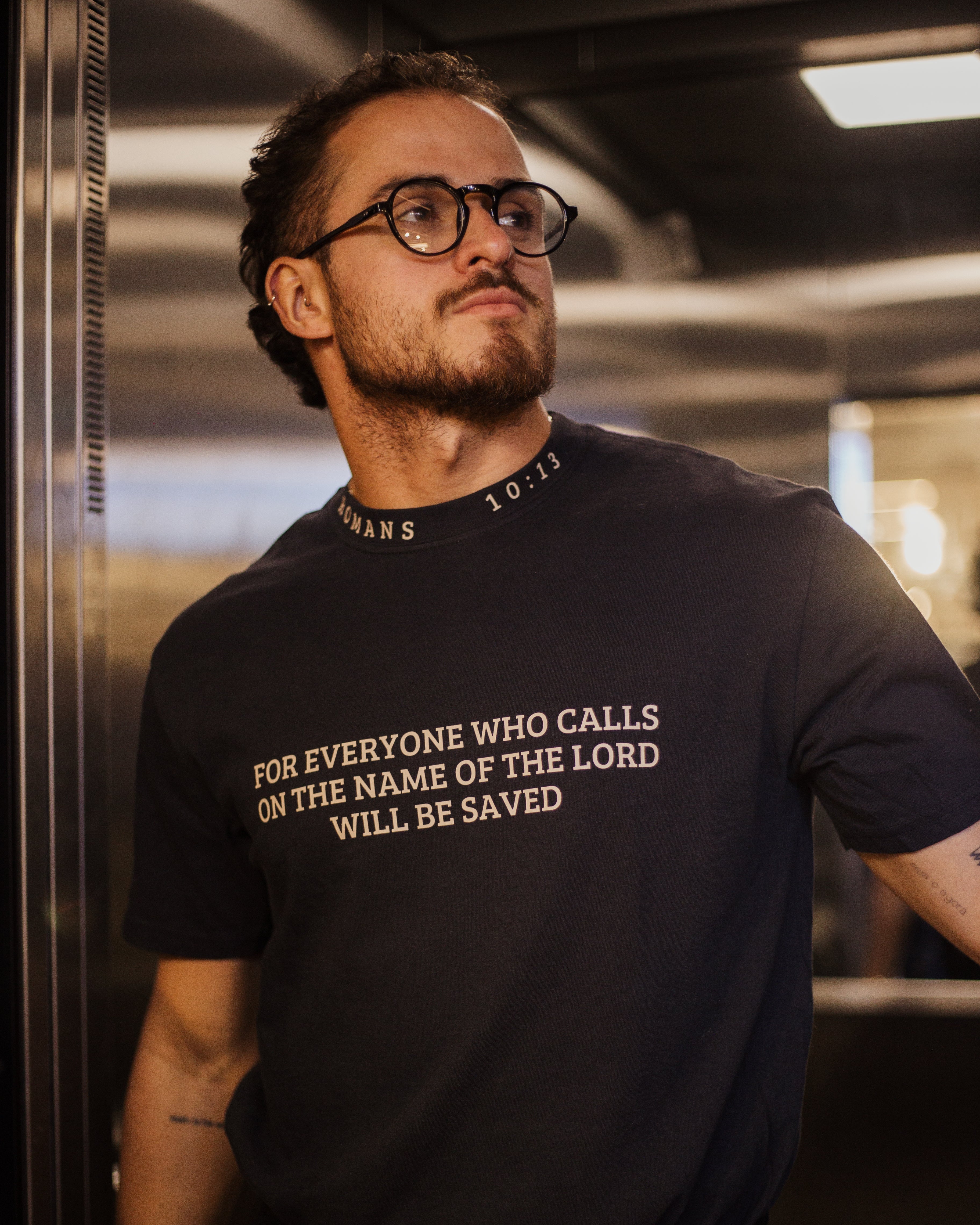A man wearing a black crew-neck t-shirt with the text 'FOR EVERYONE WHO CALLS ON THE NAME OF THE LORD WILL BE SAVED' printed in gold on the front, standing in a metallic hallway.