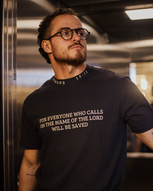 A man wearing a black crew-neck t-shirt with the text 'FOR EVERYONE WHO CALLS ON THE NAME OF THE LORD WILL BE SAVED' printed in gold on the front, standing in a metallic hallway.