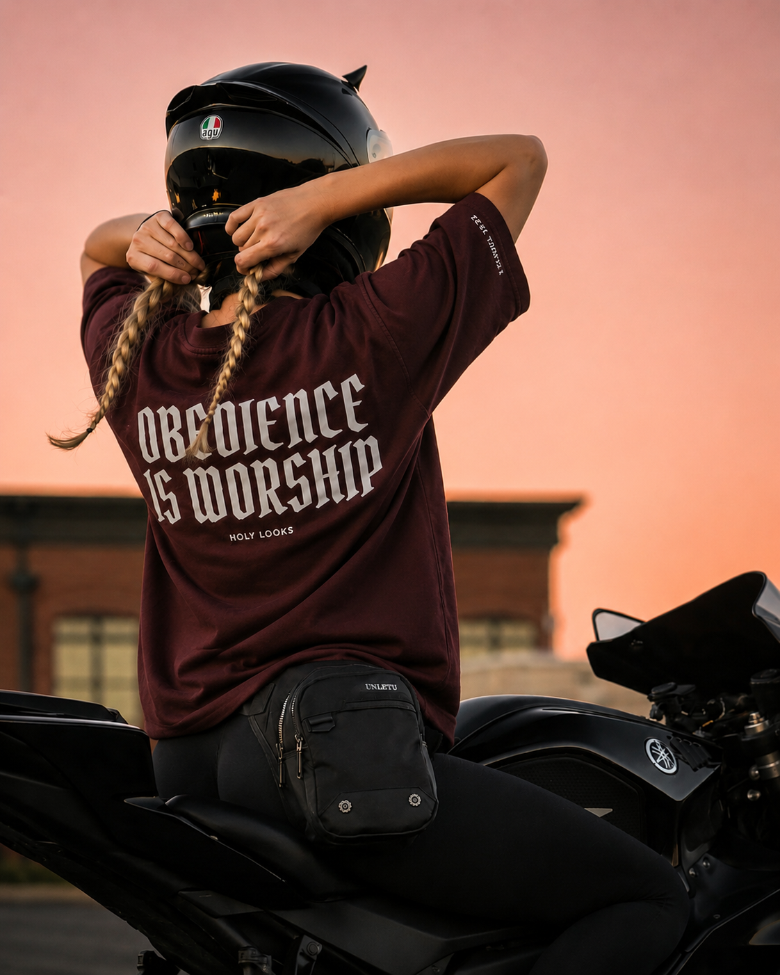 A person wearing a black motorcycle helmet and a burgundy t-shirt with the text 'OBEDIENCE IS WORSHIP' sits on a black motorcycle against a warm pink-orange sunset sky.
