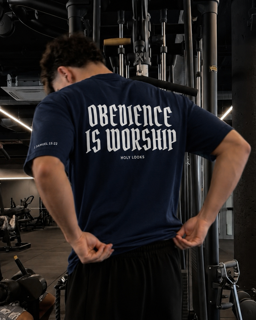 A man in a gym wearing a navy blue t-shirt with 'OBEDIENCE IS WORSHIP' printed in white on the back, paired with black sweatpants.
