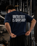 A man in a gym wearing a navy blue t-shirt with 'OBEDIENCE IS WORSHIP' printed in white on the back, paired with black sweatpants.